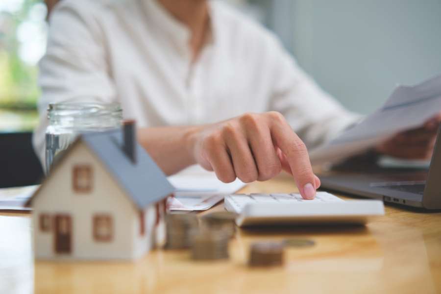 A person using a calculator next to a house model and coins to compare and choose the right insurance plan.