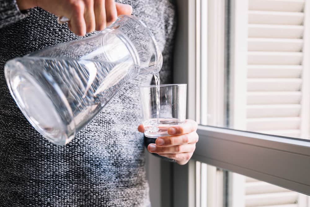 A person pouring fresh, clear water from a glass pitcher into a drinking glass by a window, highlighting superior water quality and taste.