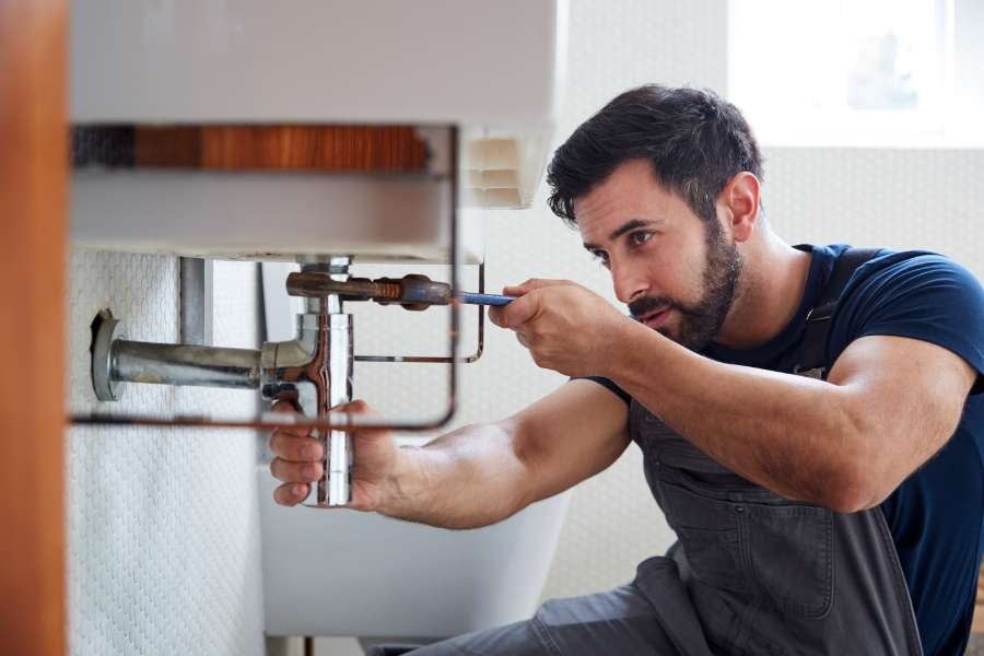 A professional plumber fixes a leaking faucet and pipes under a sink using a wrench.